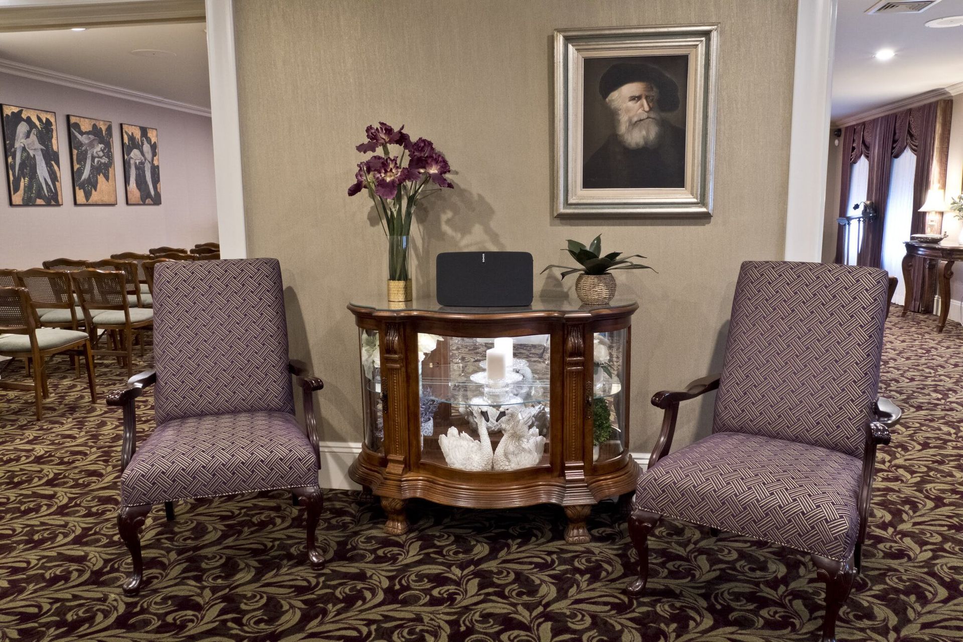 Two patterned chairs face a decorative table with flowers and a portrait in a formal reception area.