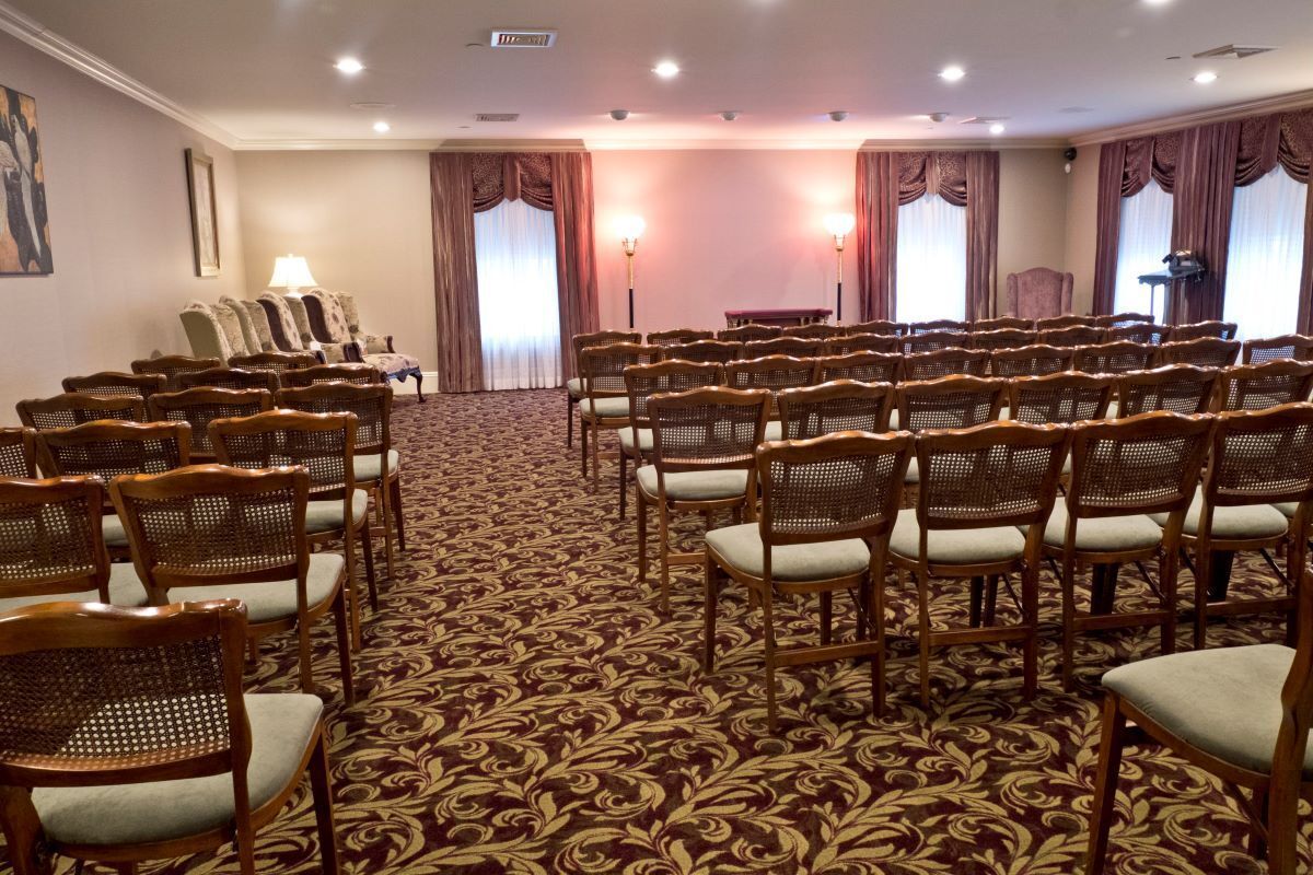 Rows of chairs face a stage in a formal room, possibly a funeral home, with patterned carpet and drapes.