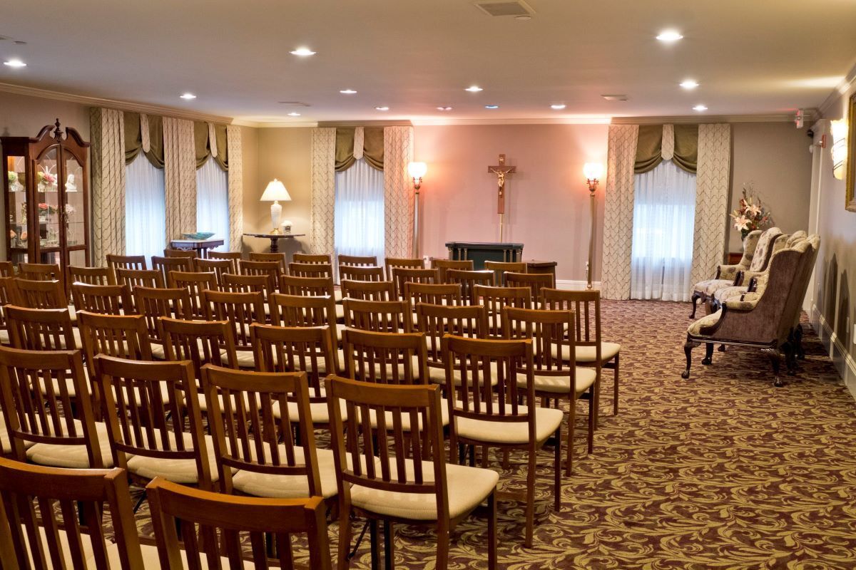 Rows of chairs face a cross in a chapel. Beige walls, carpet, and curtains, soft lighting.