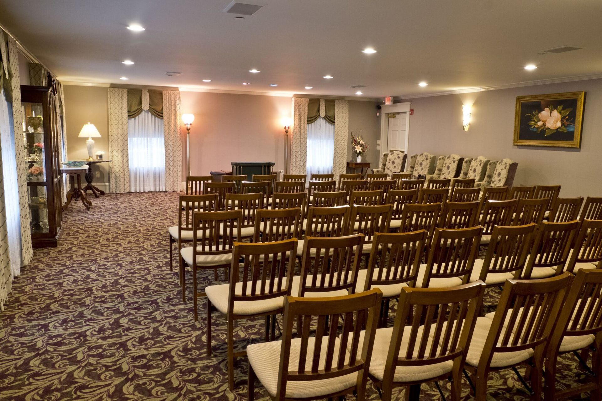 Interior of a funeral home chapel with rows of wooden chairs, patterned carpet, and artwork on the wall.