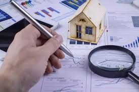 Mano con bolígrafo sobre informes financieros, maqueta de casa pequeña, lupa.