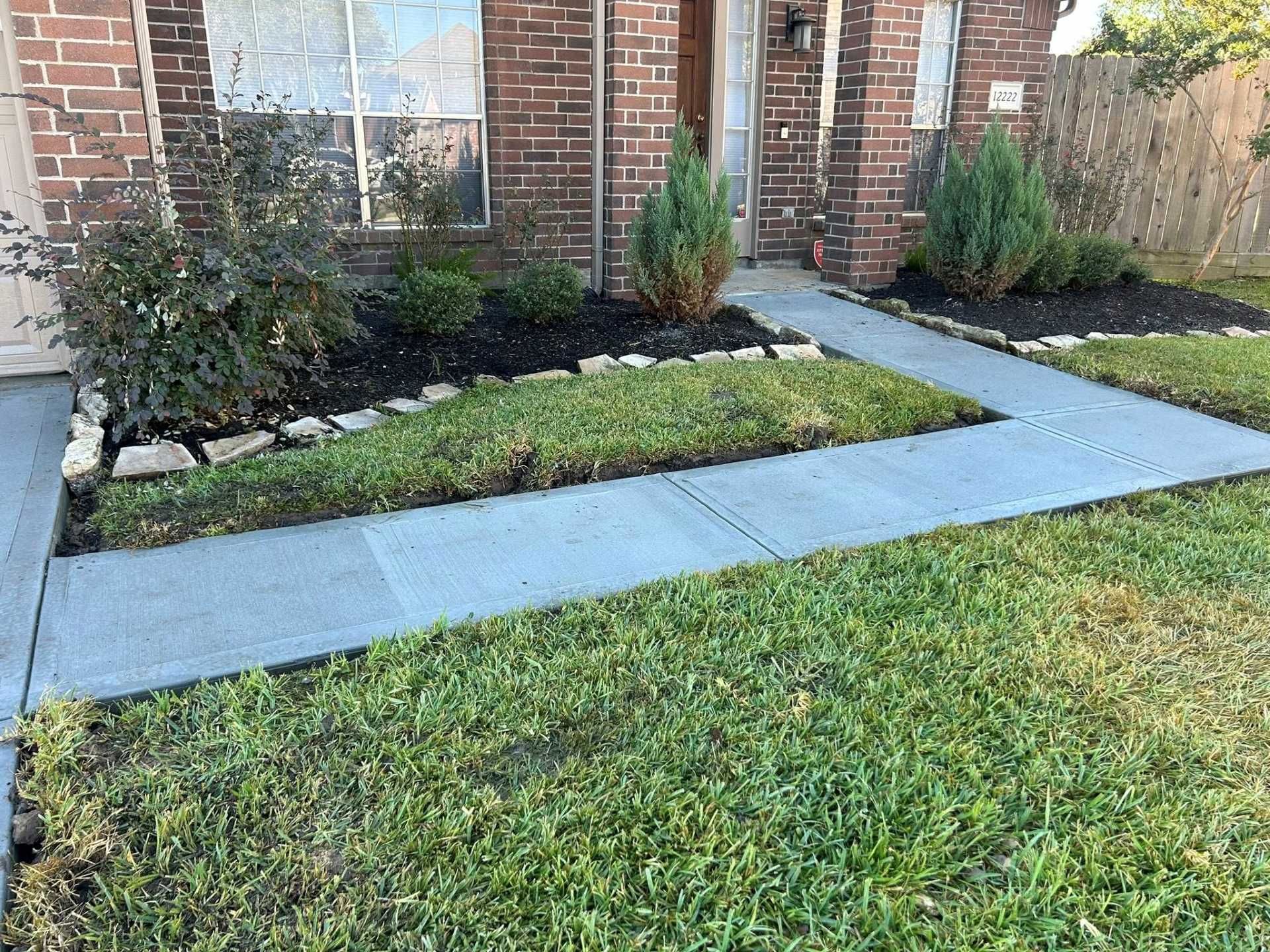 Brick house front yard with concrete walkway, grass, mulch bed, and small shrubs