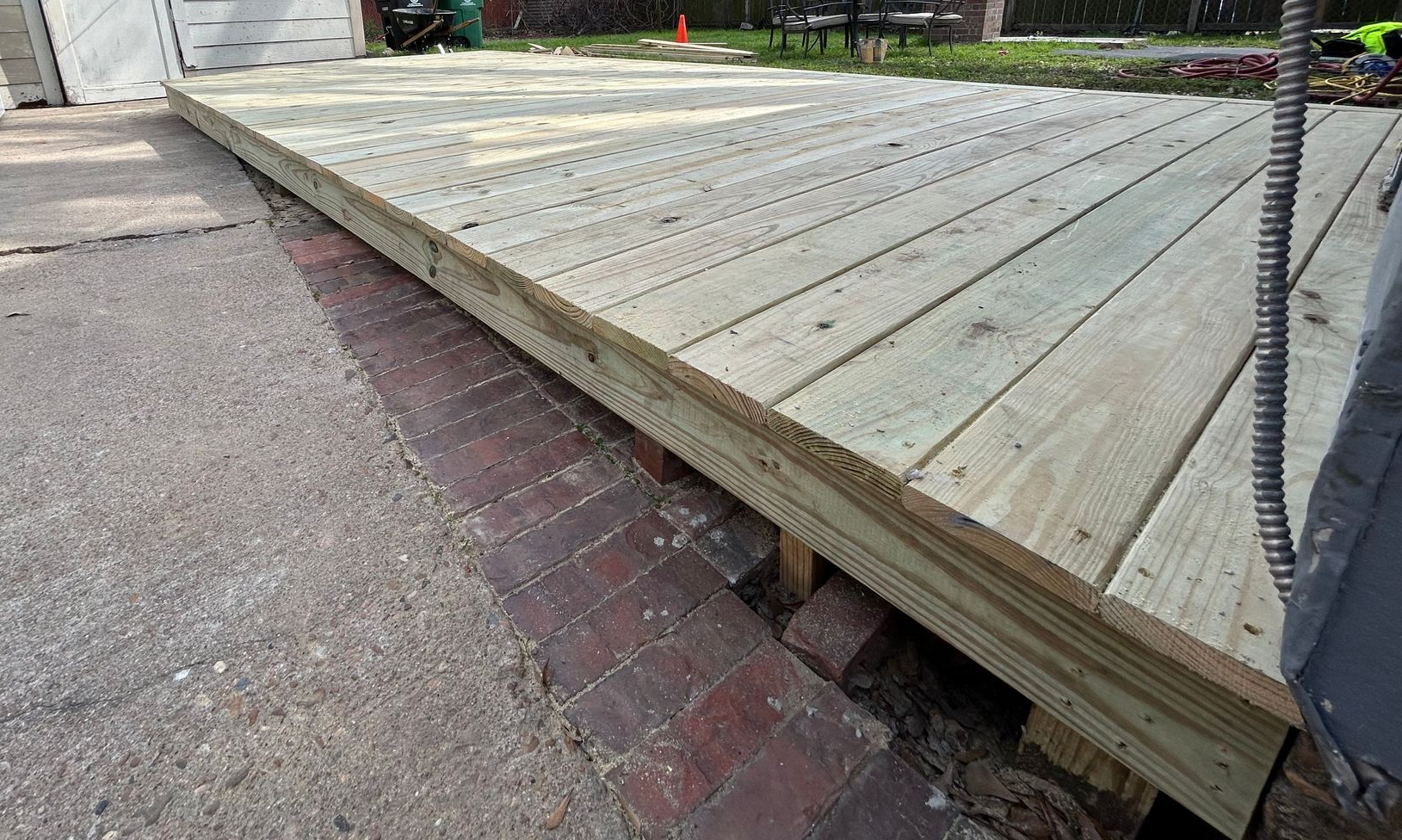 Low wooden deck or stage beside a brick and asphalt walkway, with metal posts at the corner.