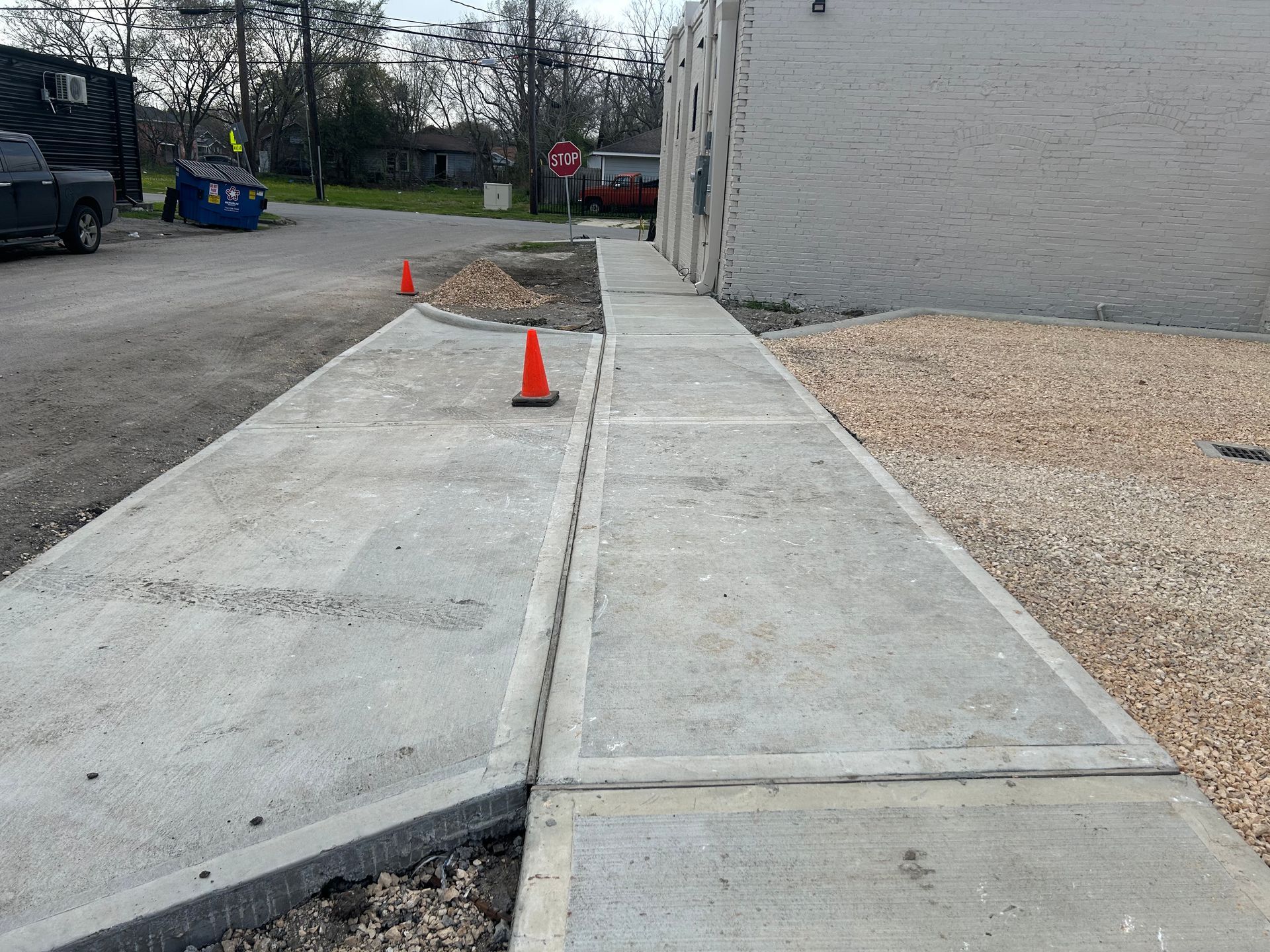Concrete sidewalk with orange cones beside a white building and gravel parking area
