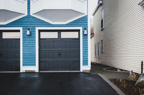 Blue house facade with two dark garage doors beside a white siding home and wet driveway