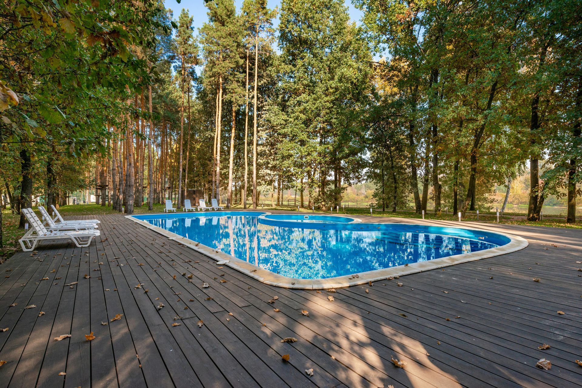 Wooden deck beside a blue outdoor pool surrounded by trees, with white lounge chairs.