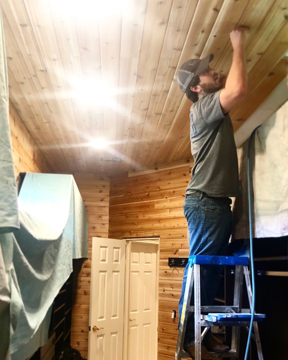 Man on ladder installing wooden trim on ceiling inside a room with wood-paneled walls.