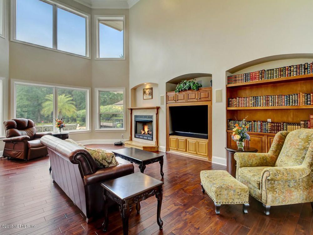 Living room with high ceilings, large windows, fireplace, built-in TV, bookcase, and brown leather furniture.