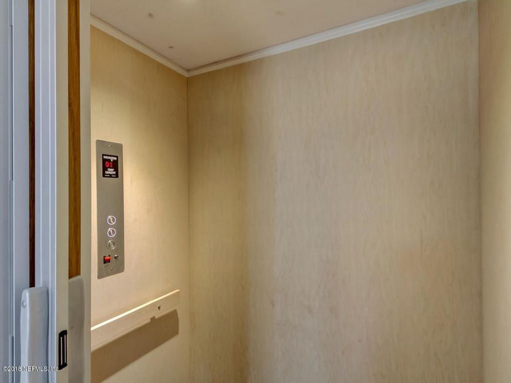 Interior of a beige elevator with call buttons and a handrail. Doorway visible on the left.
