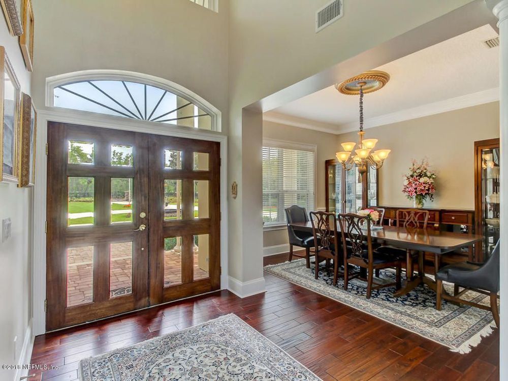 Grand foyer with double wooden doors, dining room with table, chandelier, and rug.