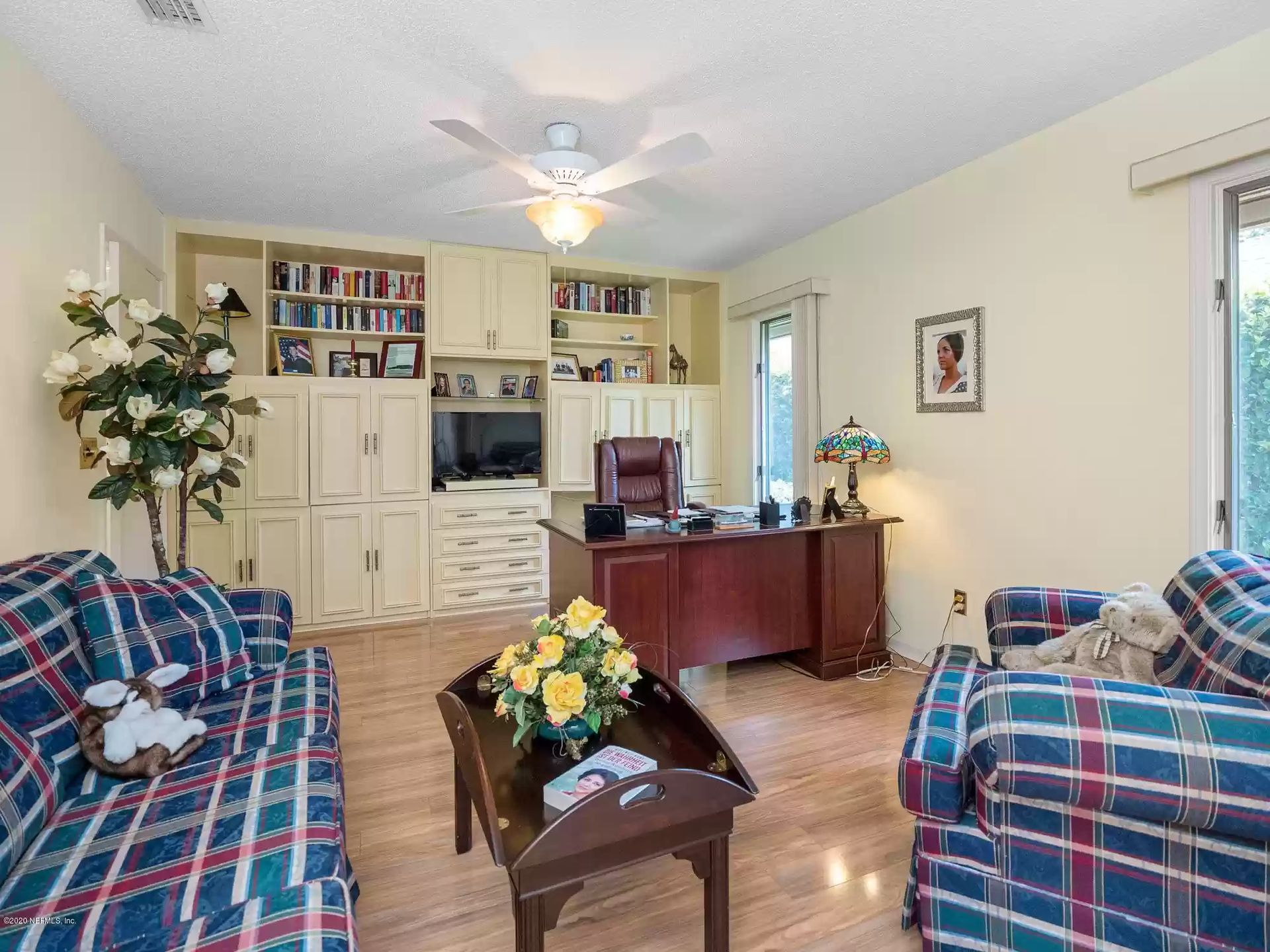 Home office with built-in cabinetry, desk, sofas, and a ceiling fan. Wooden floors.