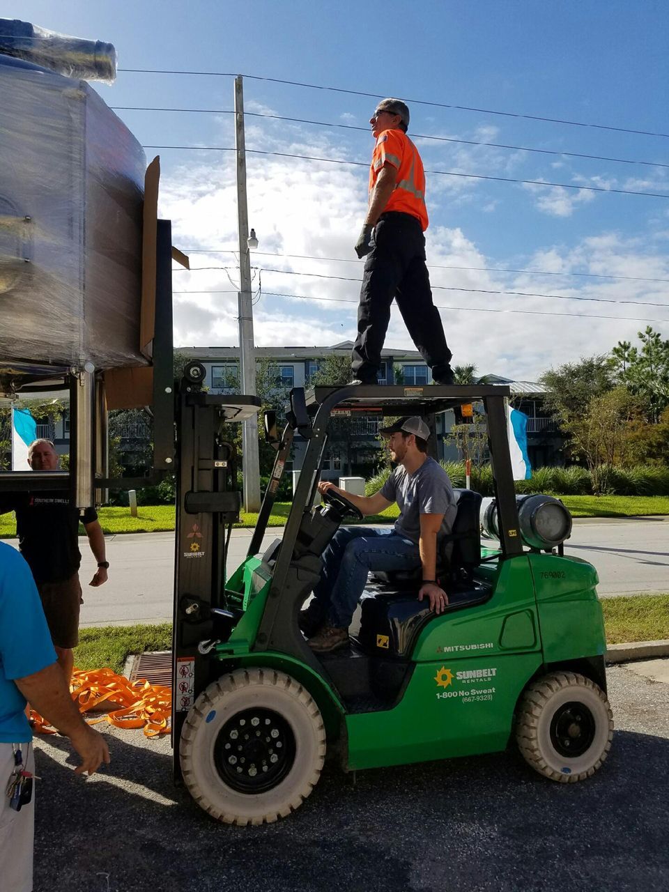 A man stands on a forklift, another drives it, aiding in moving a large object outdoors.