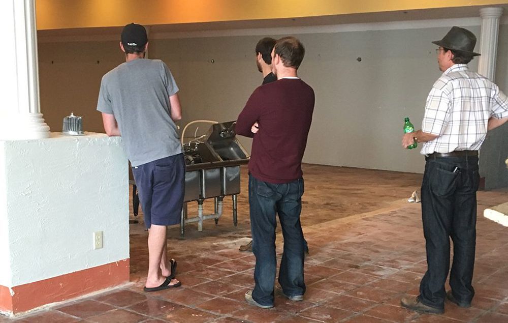 Four men in a vacant room, discussing. A stainless steel sink unit. One man with a beer.
