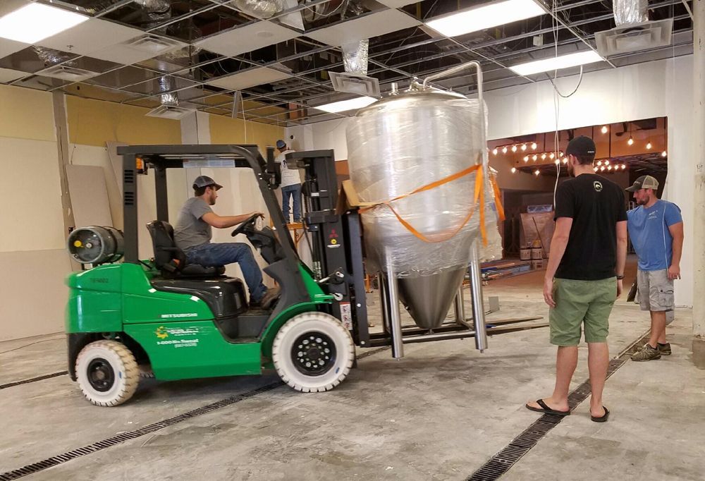 A forklift in a brewery carries a large, stainless steel tank. Two men watch.