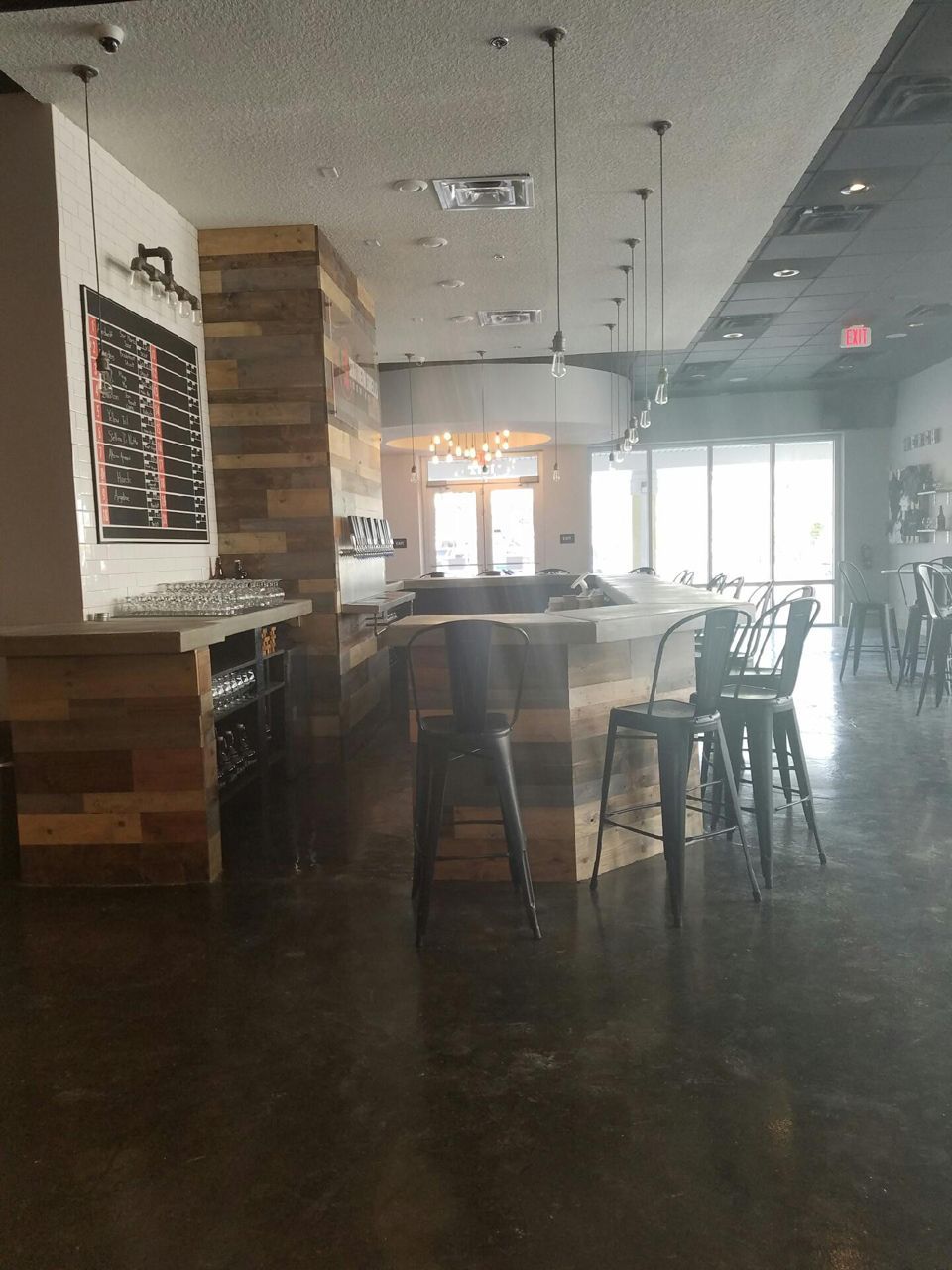 Empty bar with wood-paneled counter and stools, dark floor, bright windows, and a sign.