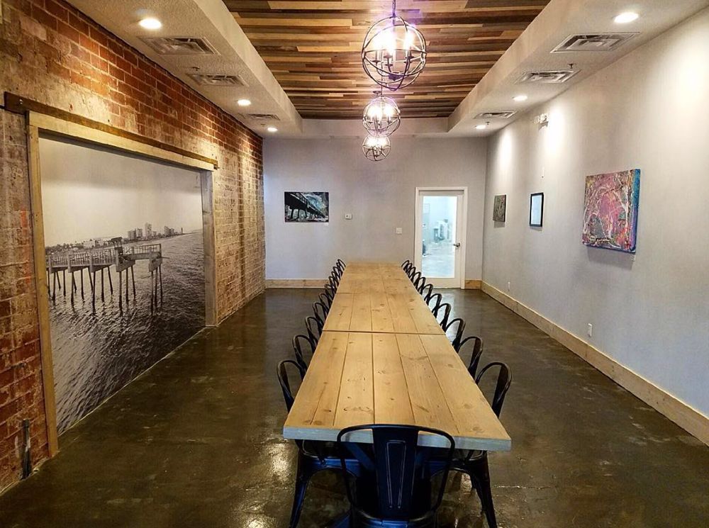 Long wooden table with black chairs in an art gallery; exposed brick wall, art prints, and a photograph.