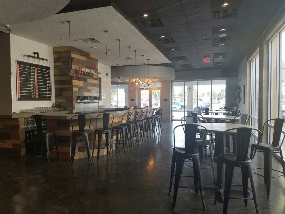 Interior of a bar with a long counter, bar stools, tables, and large windows. Dark floor, exposed beams, neutral colors.