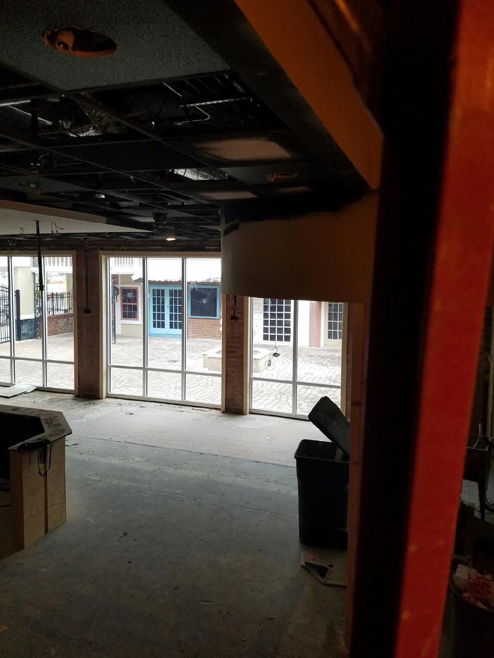 Interior construction site with large windows, exposed ceiling, and trash bin.