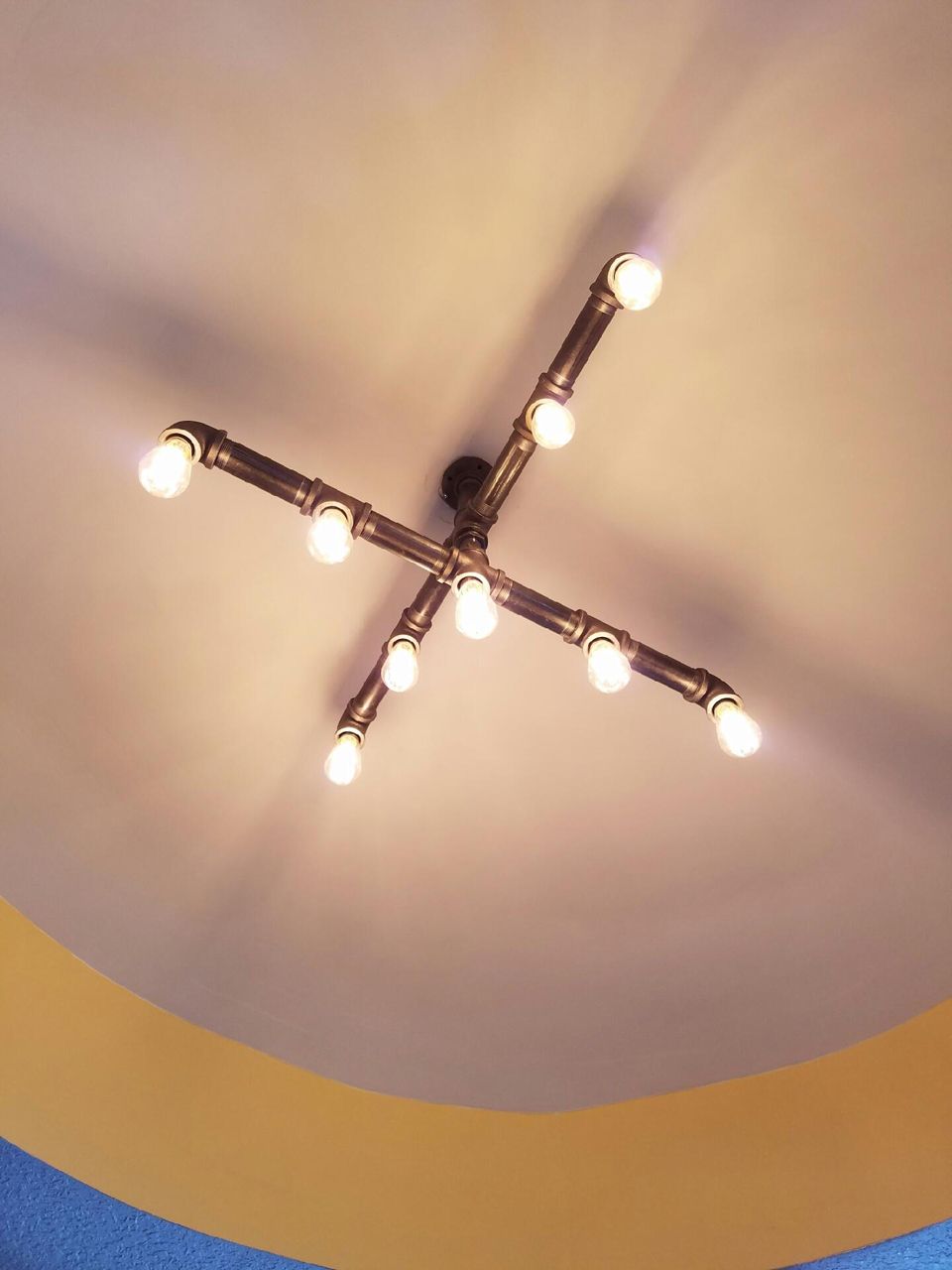 A ceiling-mounted light fixture made of bronze pipes in an X-shape, illuminating a cream-colored ceiling.