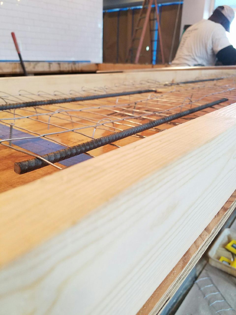 Wooden formwork with rebar and wire mesh, construction worker in the background.