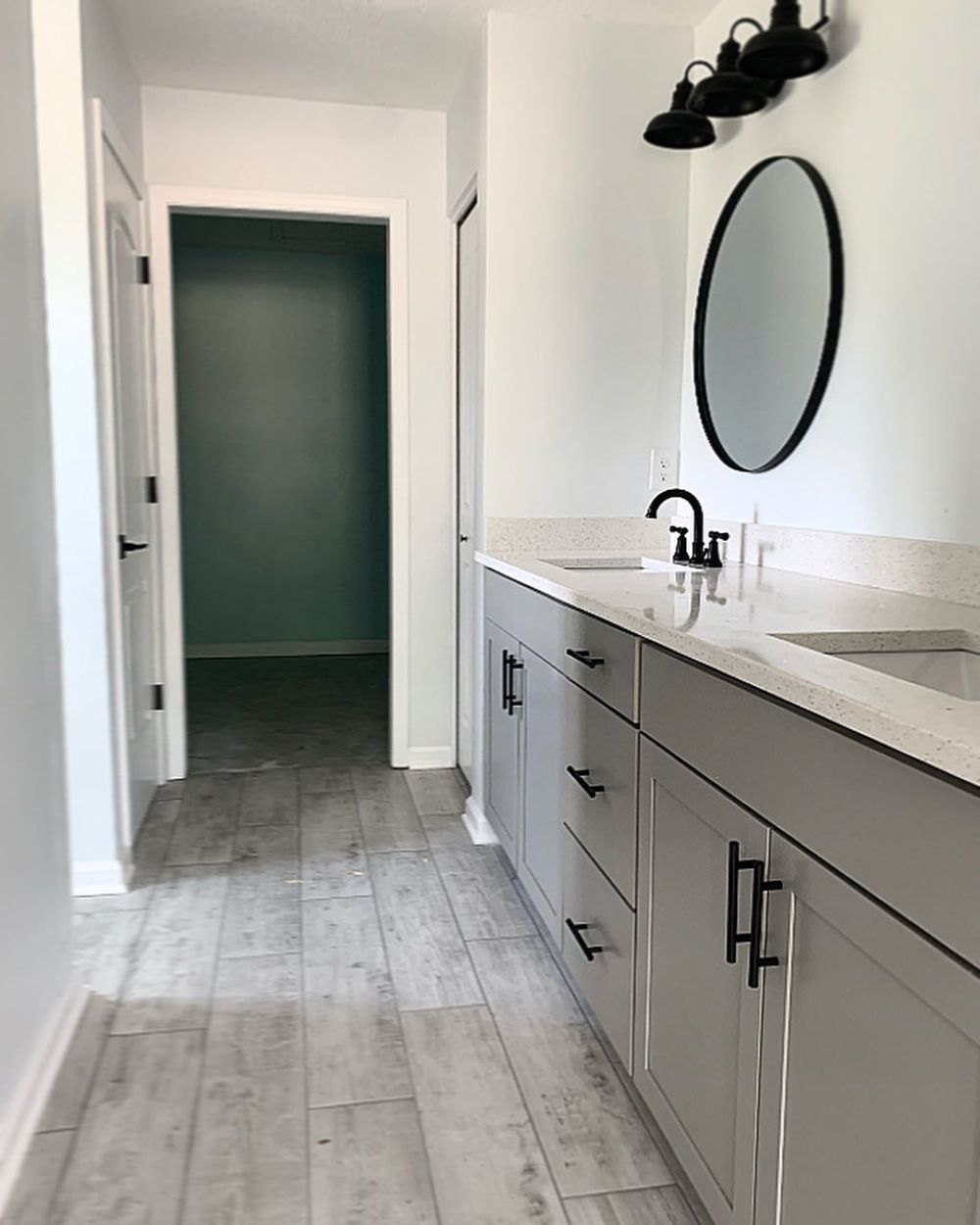 Modern bathroom with gray cabinets, black fixtures, oval mirror, and light gray flooring.