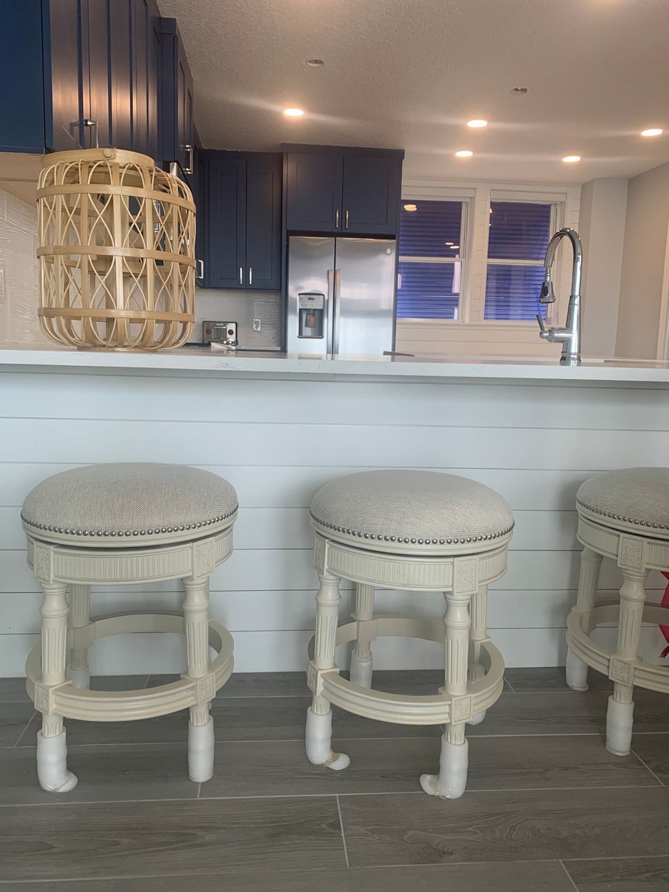 Three white bar stools in front of a kitchen island. Navy cabinets, stainless steel fridge, and a woven light fixture are in the background.