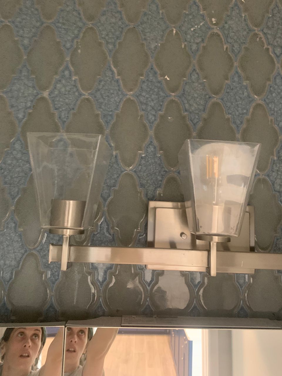 Woman cleaning a bathroom mirror, light fixture with two sconces against patterned tile backdrop.
