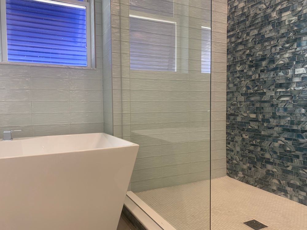 Modern bathroom with white tub, glass shower, mosaic accent wall, and blue window blinds.