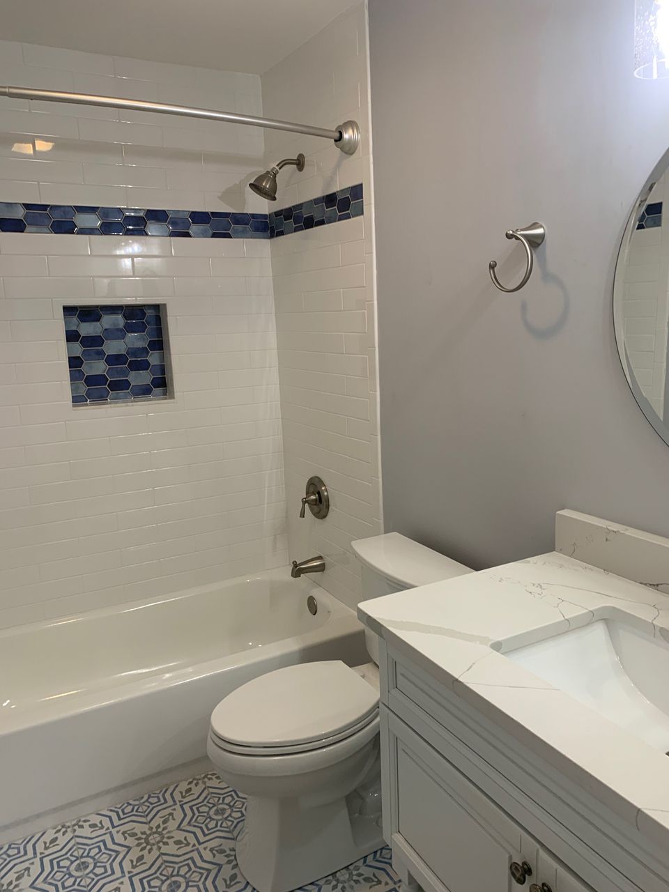 Bathroom with white tub, toilet, and vanity. Blue accent tiles and patterned floor. Round mirror.