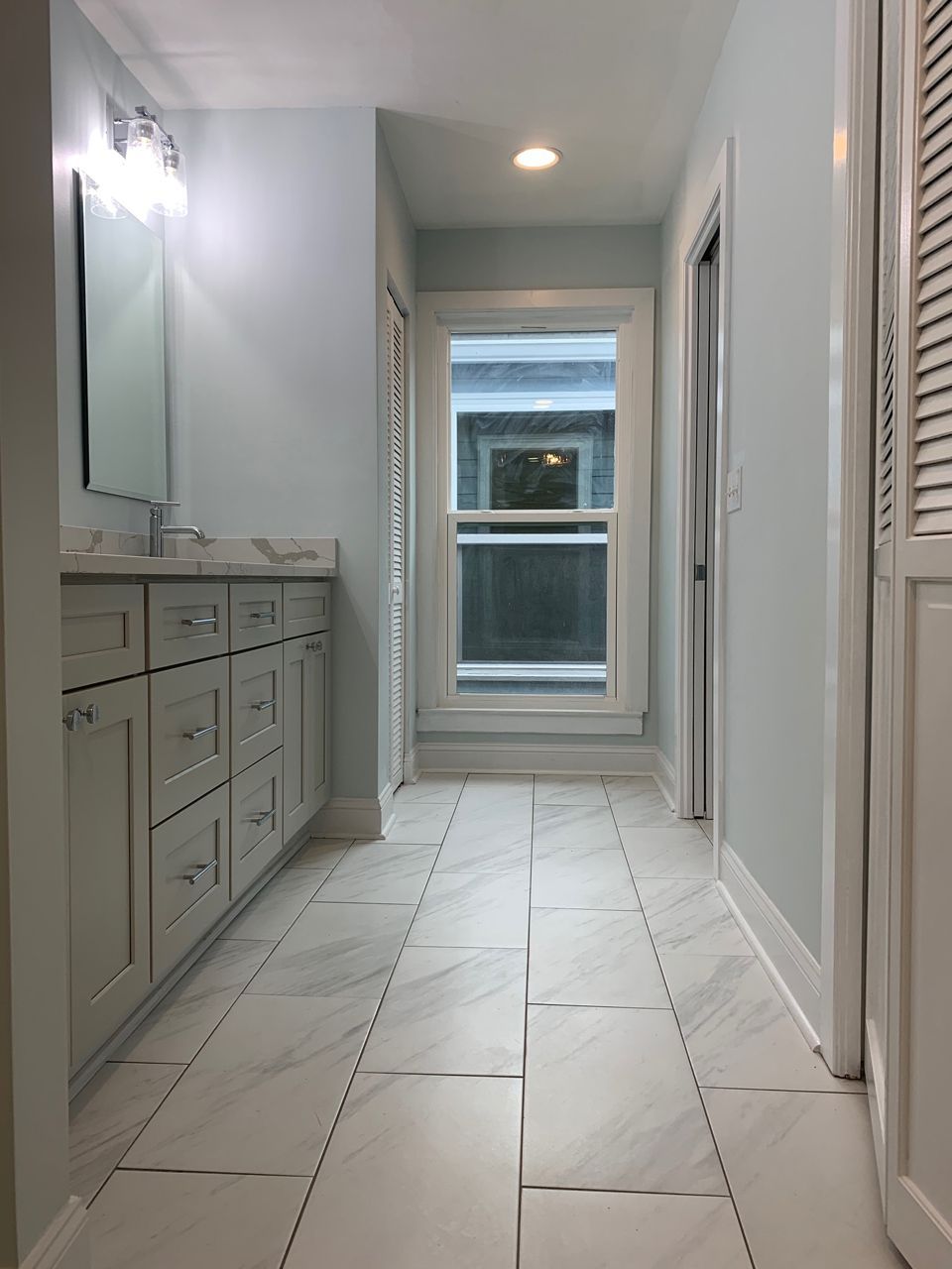 Bathroom with light blue walls, white vanity, tile floor, and a window at the end.