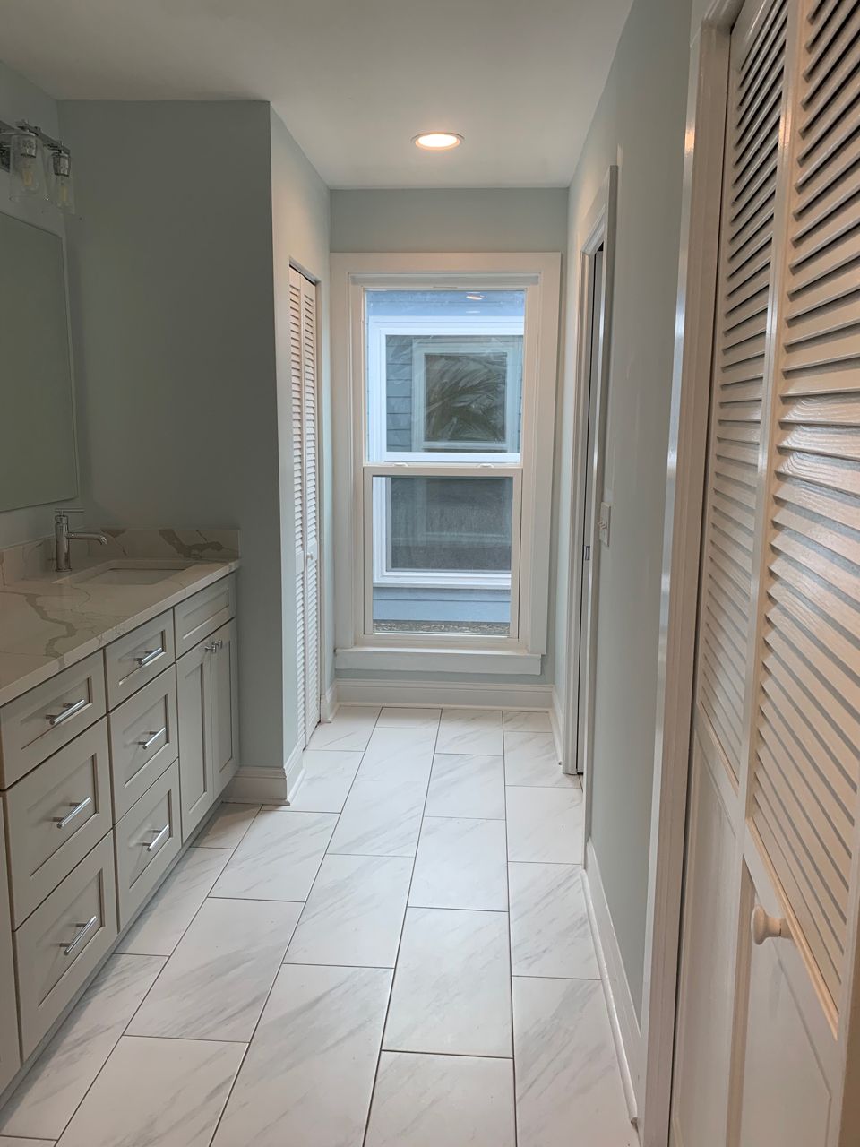 A long, narrow bathroom with light blue walls, white cabinets, and a window at the end.