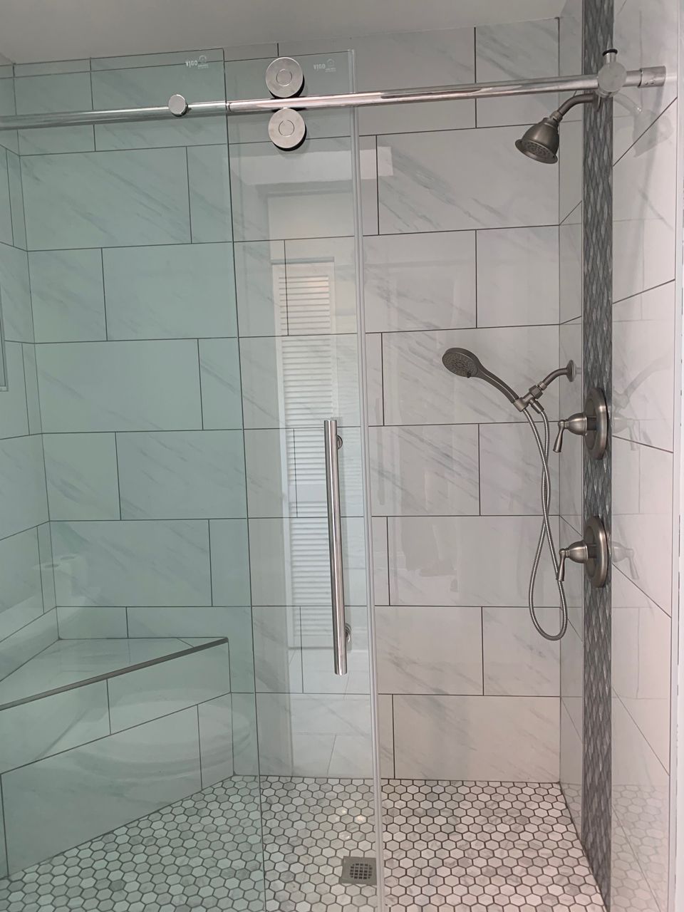 A modern bathroom shower with a clear glass door, white tiles, and a handheld showerhead.