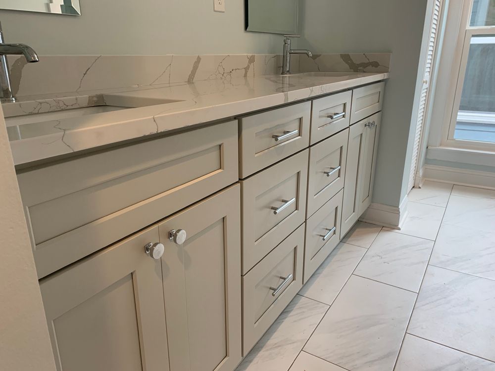 Light gray bathroom vanity with marble countertop and stainless steel hardware. White tiled floor and window.
