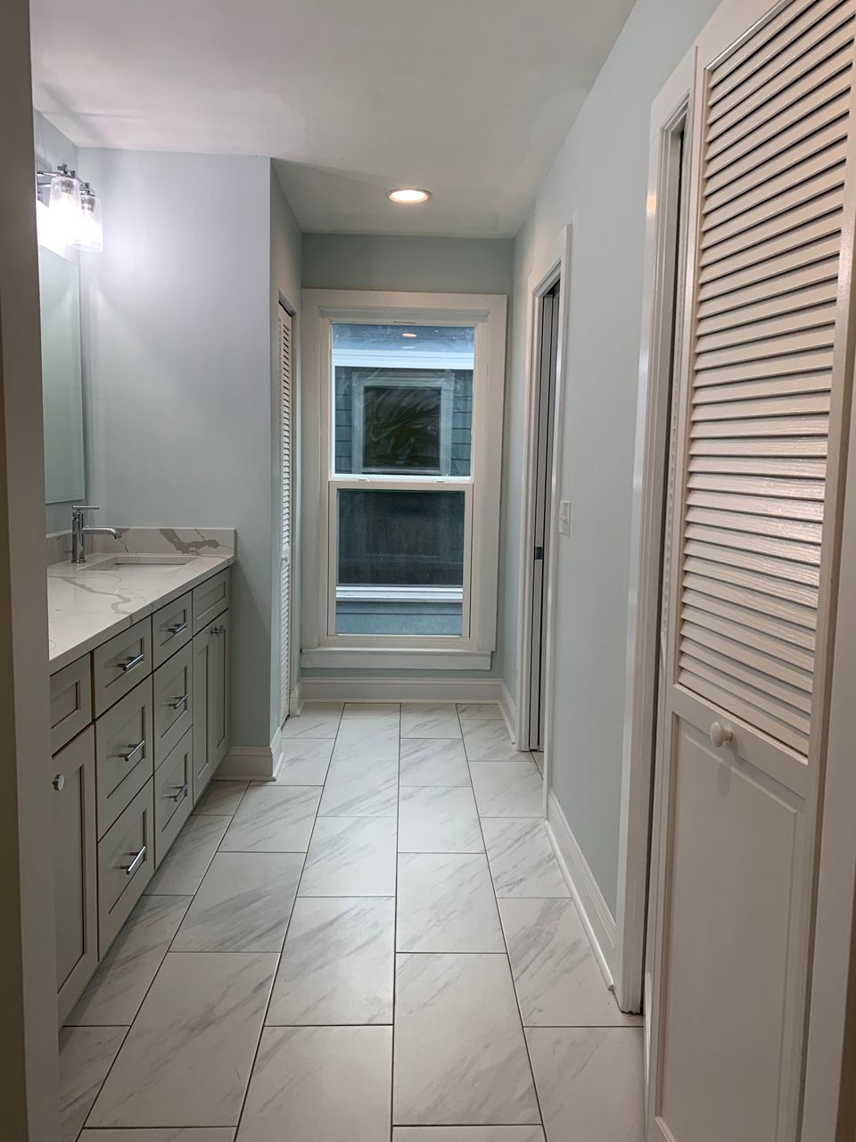 Bathroom with marble-look tile floor, pale blue walls, white cabinets, a window, and a closed shuttered door.
