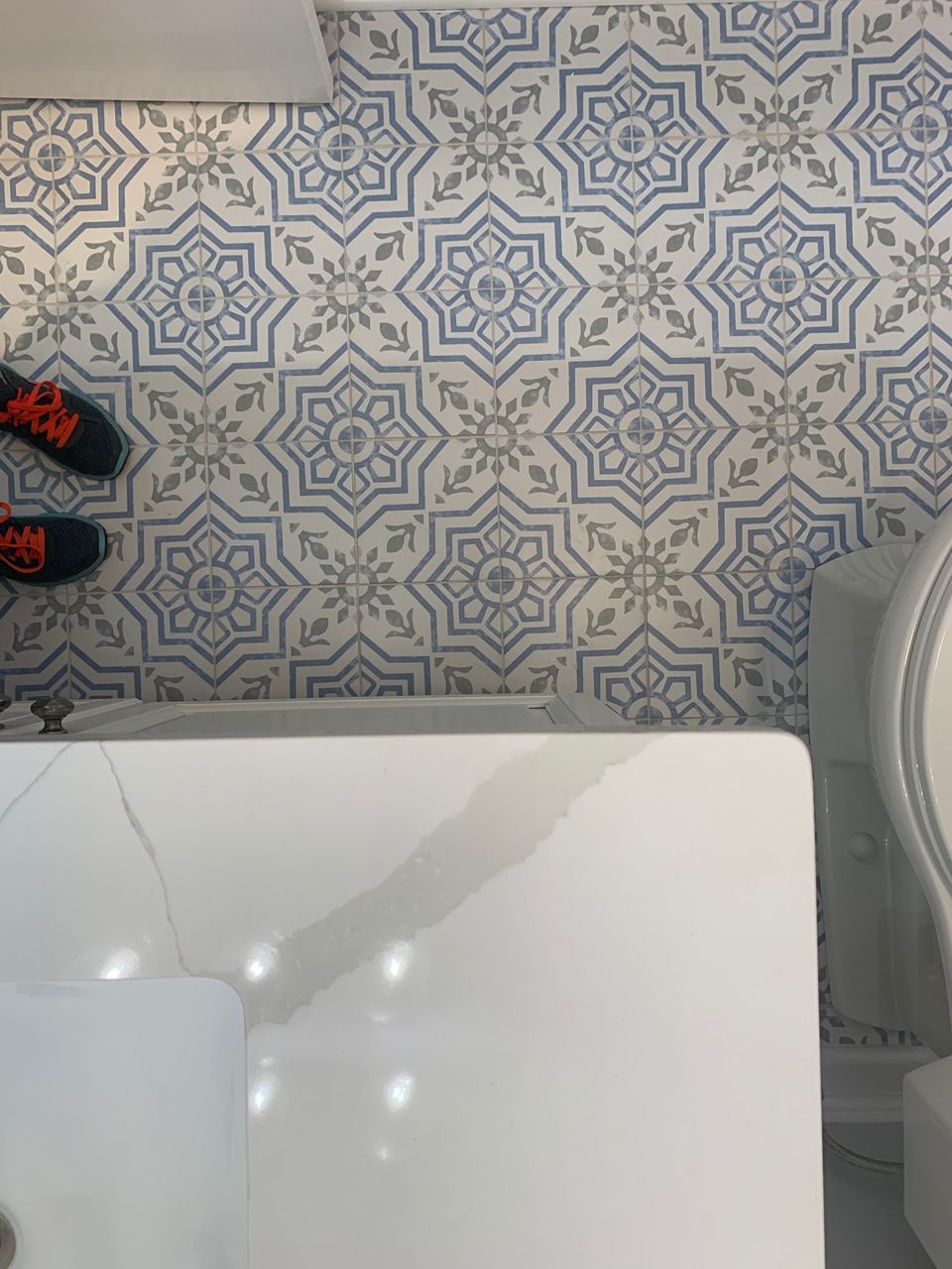 Blue and white patterned tile floor in a bathroom. White countertop and a person's feet with black and orange shoes visible.