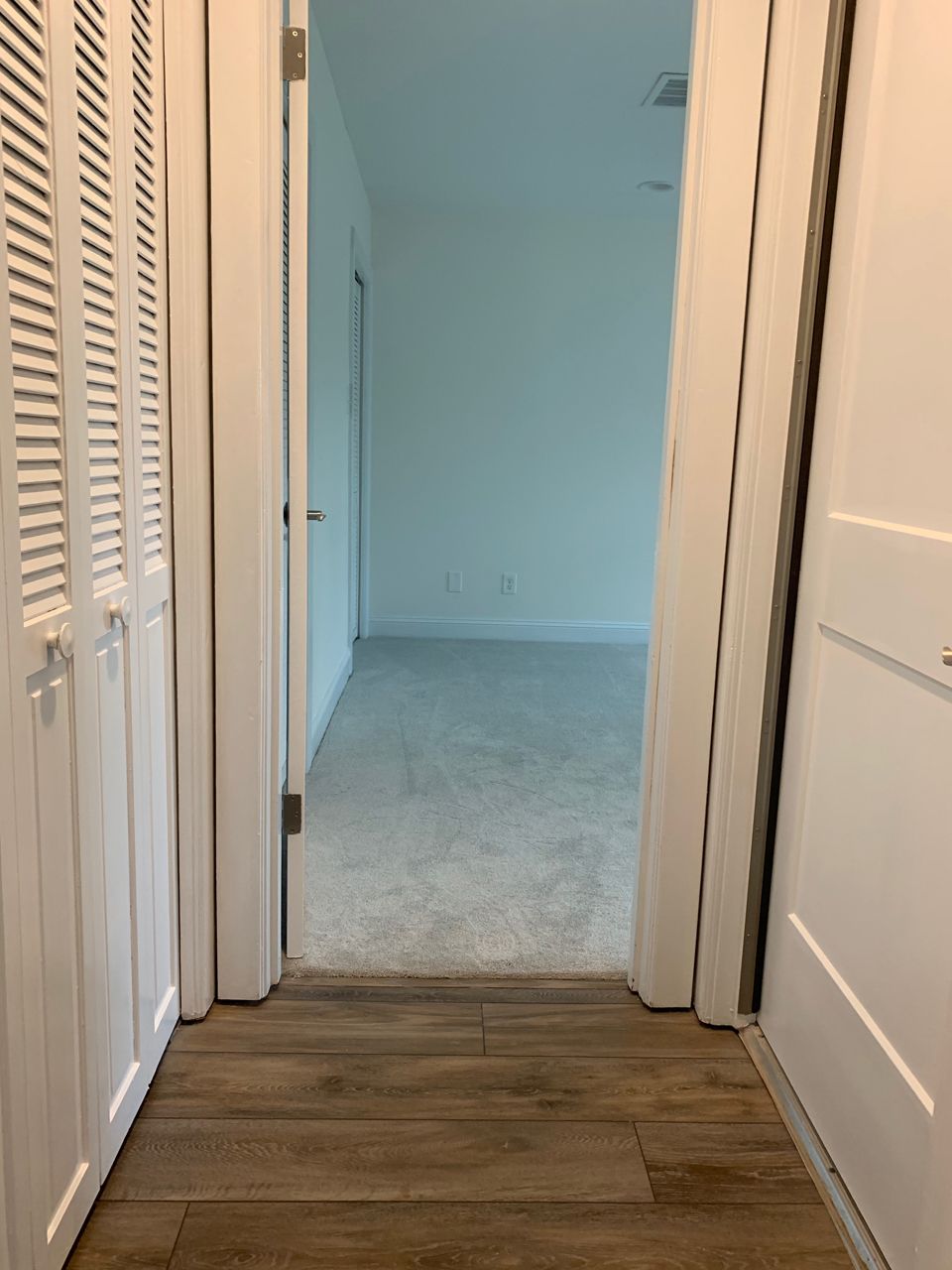 Hallway with wooden floor, two white doors, and a doorway leading to a room with gray carpet and white walls.