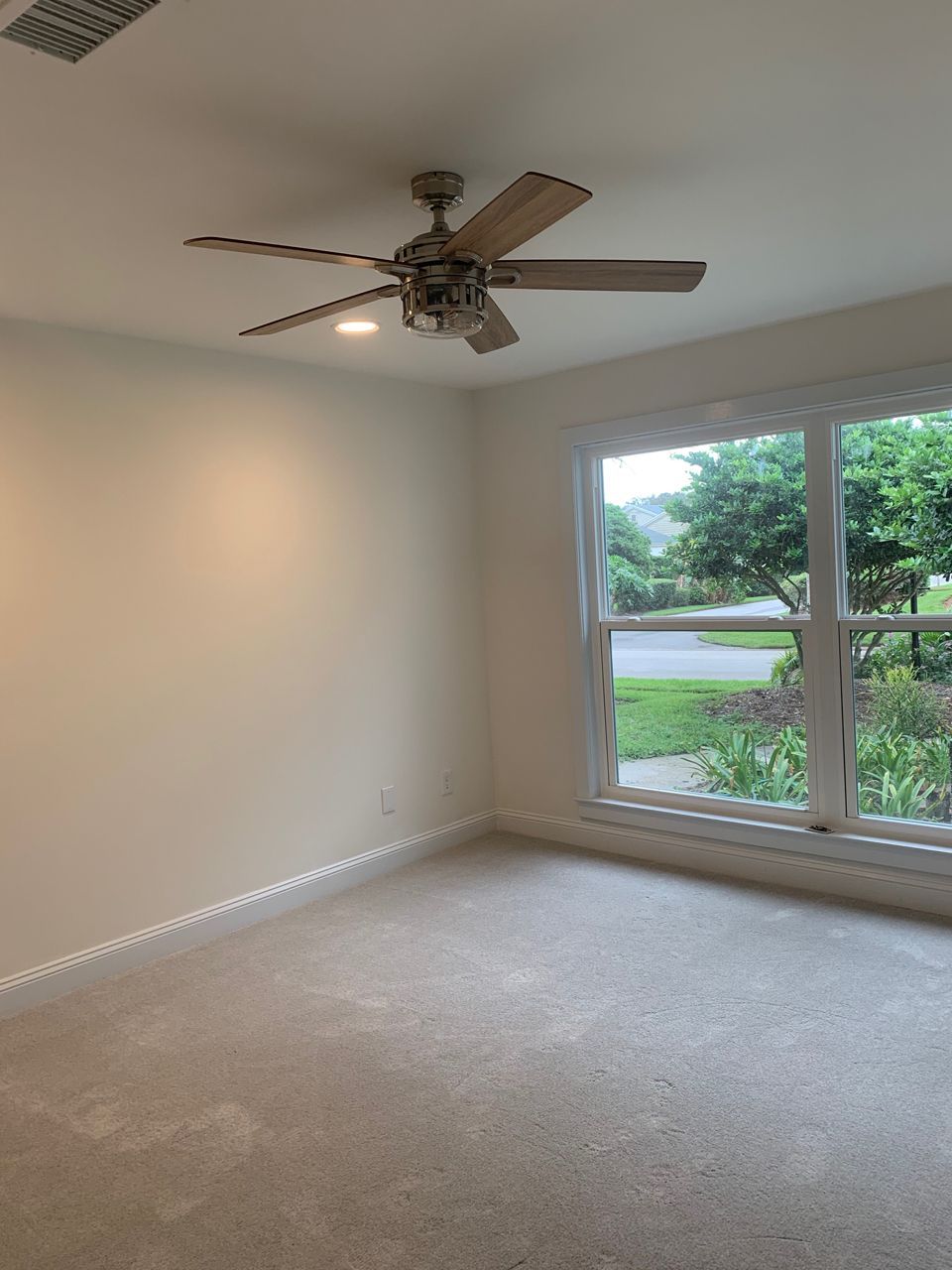 Empty room with neutral-colored walls, carpet, a ceiling fan, and a window overlooking greenery.