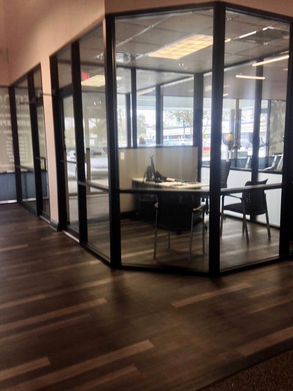 Offices with glass walls, black frames, and dark wooden floors. A desk and chairs are inside.