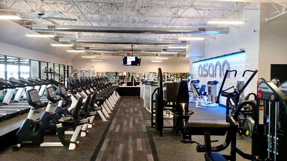 Inside a modern gym, rows of exercise machines line the floor, bright lighting.