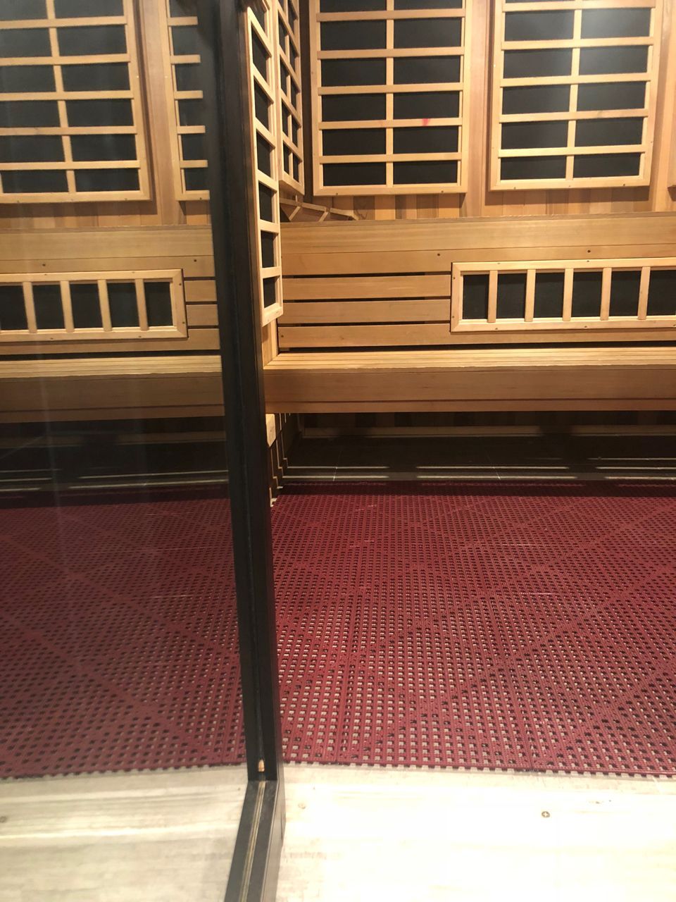 Red-floored sauna interior with wooden benches and multiple rectangular windows, seen through a glass door.