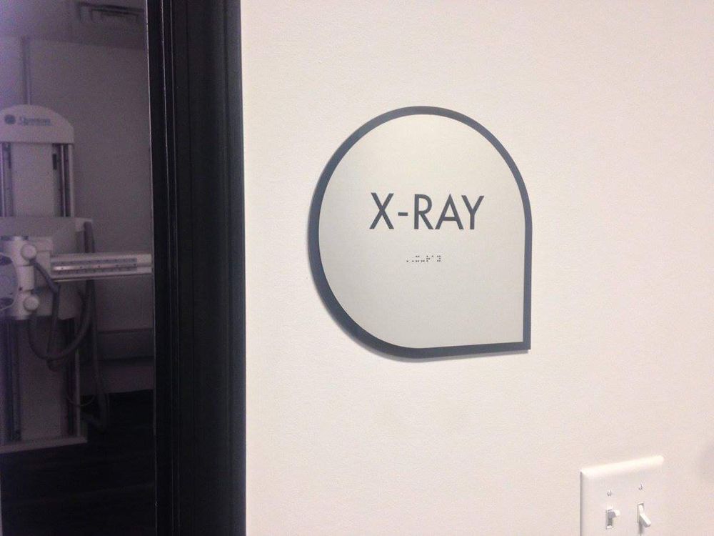 X-ray room sign with Braille on a white wall; X-ray machine visible through doorway.