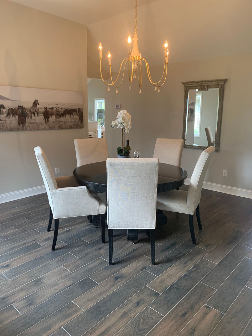 Dining room with round dark table, six cream chairs, chandelier, mirror, and artwork; wood-look tile floor.