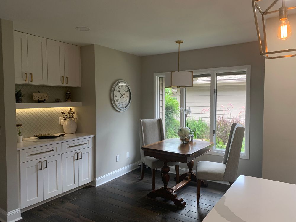 Dining area with built-in white cabinets, round clock, wooden table, two chairs by window.