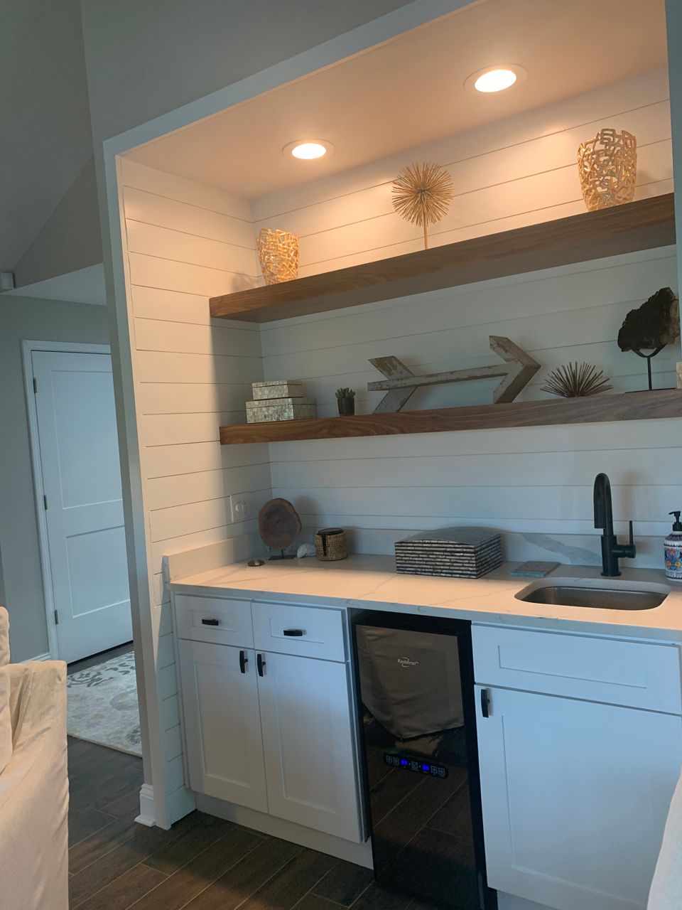 Built-in white cabinet with shelves, a sink, and mini-fridge. Decorated with gold accents on a white shiplap wall.