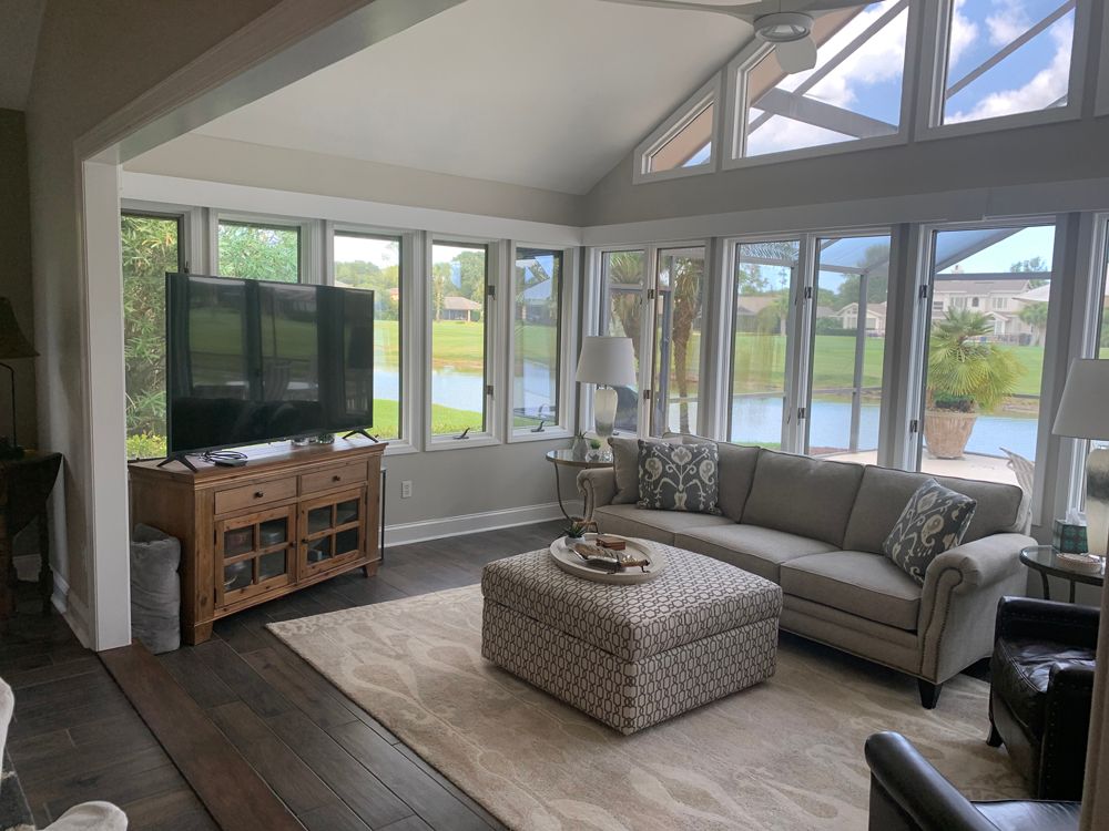Sunroom with large windows overlooking water; sofa, TV, and patterned ottoman.