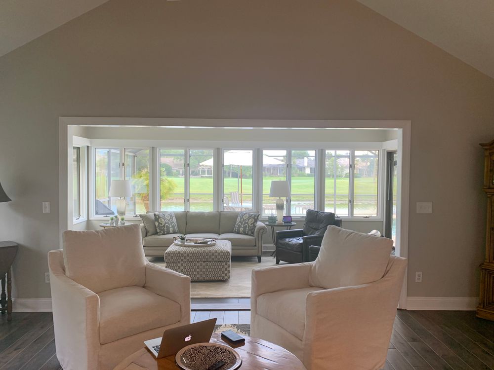 Two white armchairs face a light-filled sunroom with a sofa and windows overlooking a green lawn.