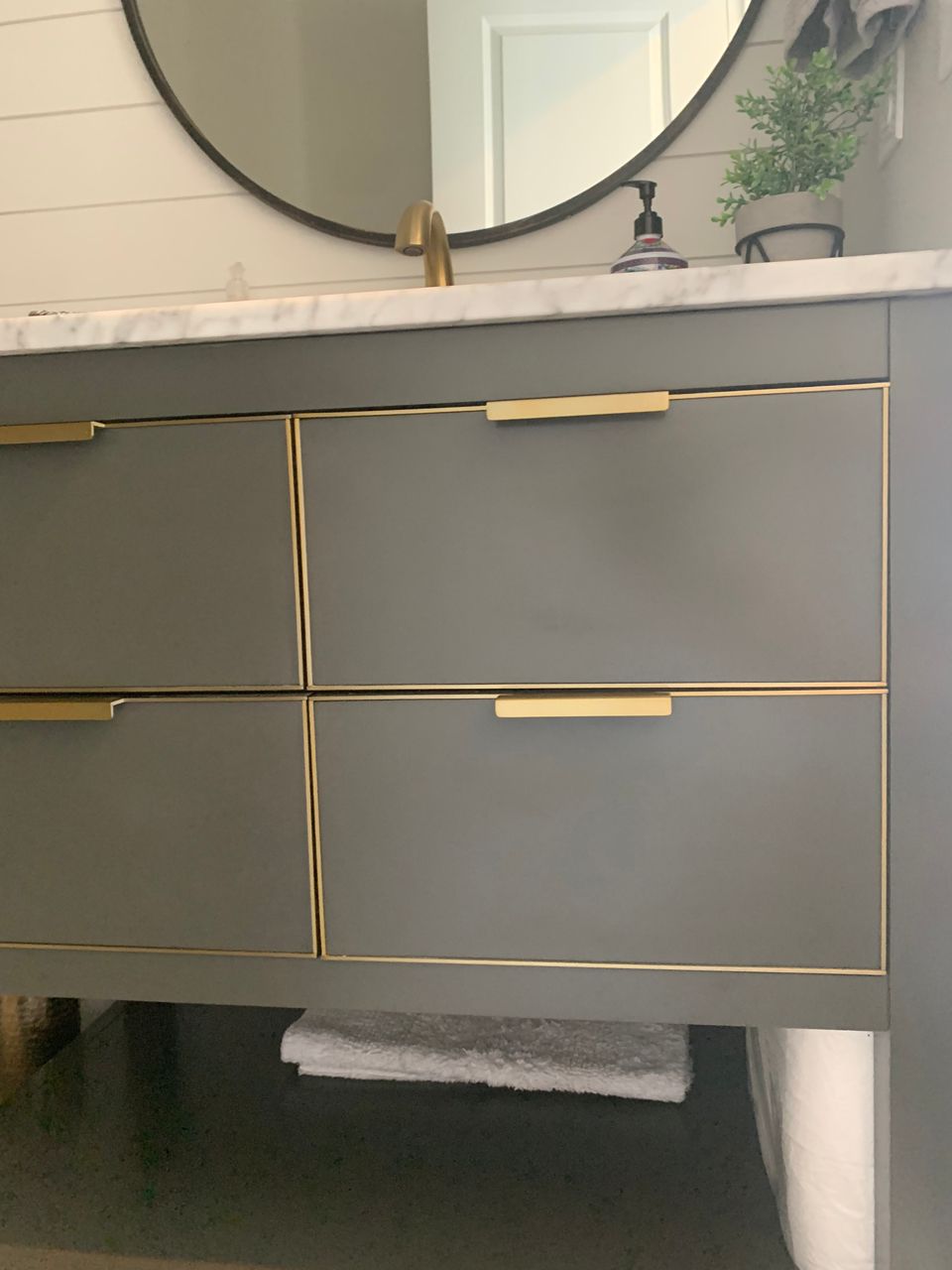 Gray bathroom vanity with gold trim and handles, marble countertop, and round mirror.