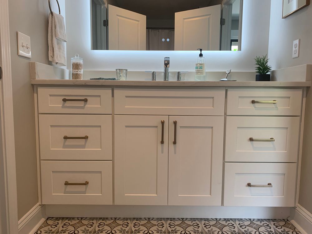 White bathroom vanity with drawers and doors, gold hardware, lit mirror, and patterned tile.
