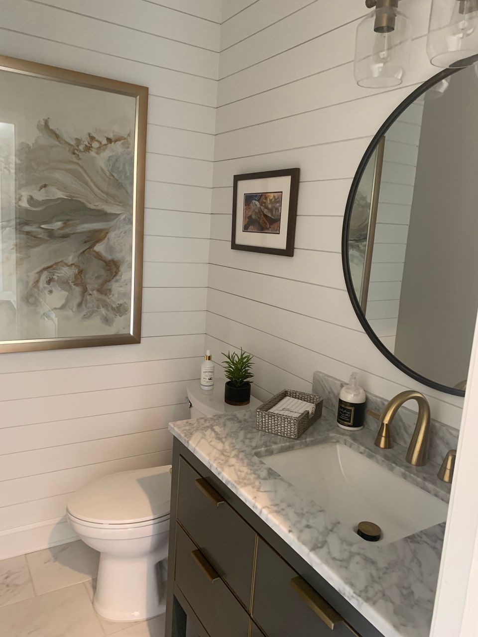 Small bathroom with white shiplap walls, gray vanity, gold fixtures, a round mirror, and artwork.