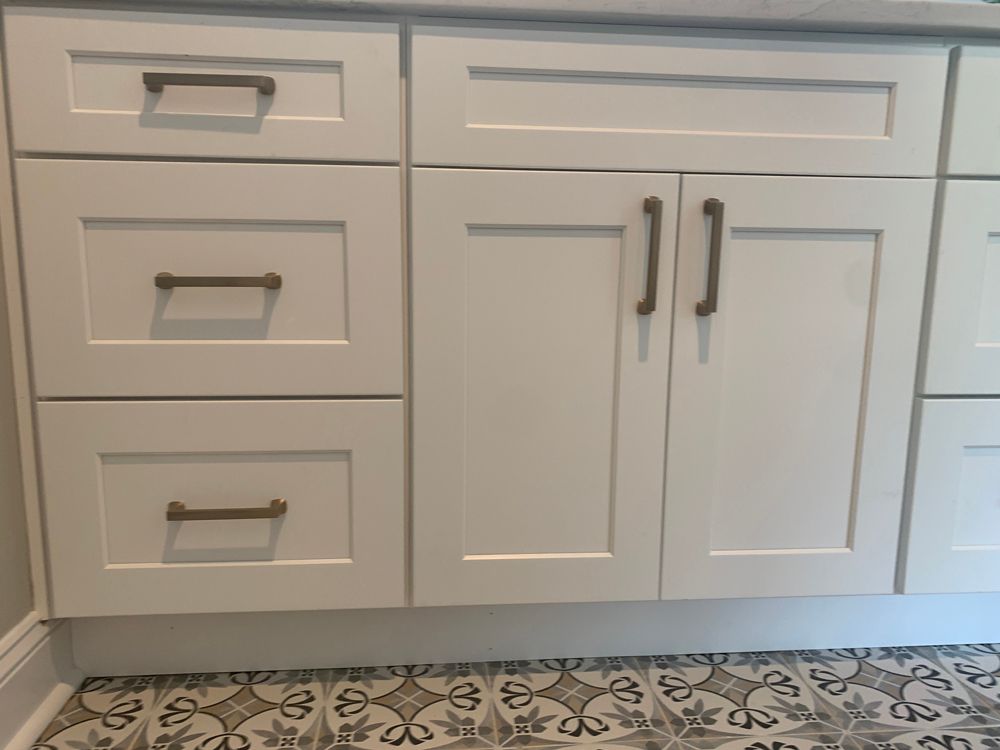 White bathroom vanity with drawers and cabinets, gold hardware. Patterned floor tile.
